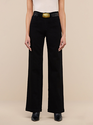 Wearing the Scottie Wide-Leg Jean in black premium stretch denim, a white sleeveless top, a black textured belt with a large gold buckle, and black pointed-toe shoes, the person stands against a beige background.
