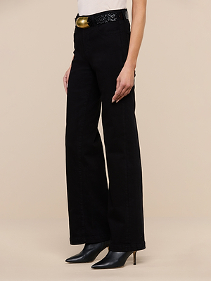 A person wearing the Scottie Wide-Leg Jean in premium stretch denim, styled with a white top, a black textured belt with a gold buckle, and black pointed-toe heeled boots, stands against a beige background.