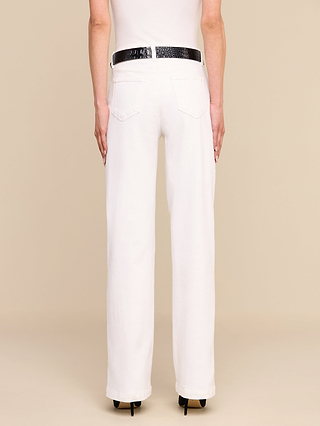Una persona aparece de espaldas, vestida con una camiseta blanca sin mangas y unos vaqueros Scottie Wide-Leg de color blanco, ceñidos con un cinturón negro y combinados con zapatos de tacón negros, sobre un fondo beige.