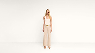 A woman wearing sunglasses, a white sleeveless top, beige wide-leg pants, and tan heels stands against a plain white background.