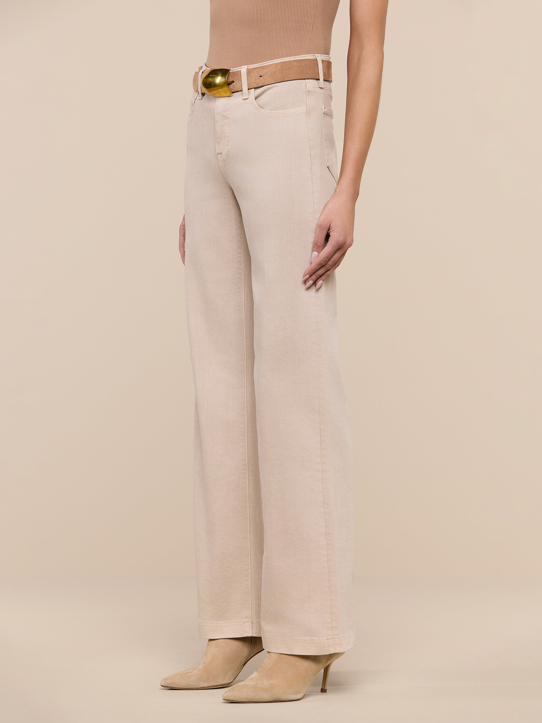 A person wearing the Scottie Wide-Leg Jean in beige—crafted from premium stretch denim and featuring a high-rise, wide-leg silhouette—is styled with a matching beige belt and tan pointed-toe heels, standing against a plain white background. The upper body and head are not visible.