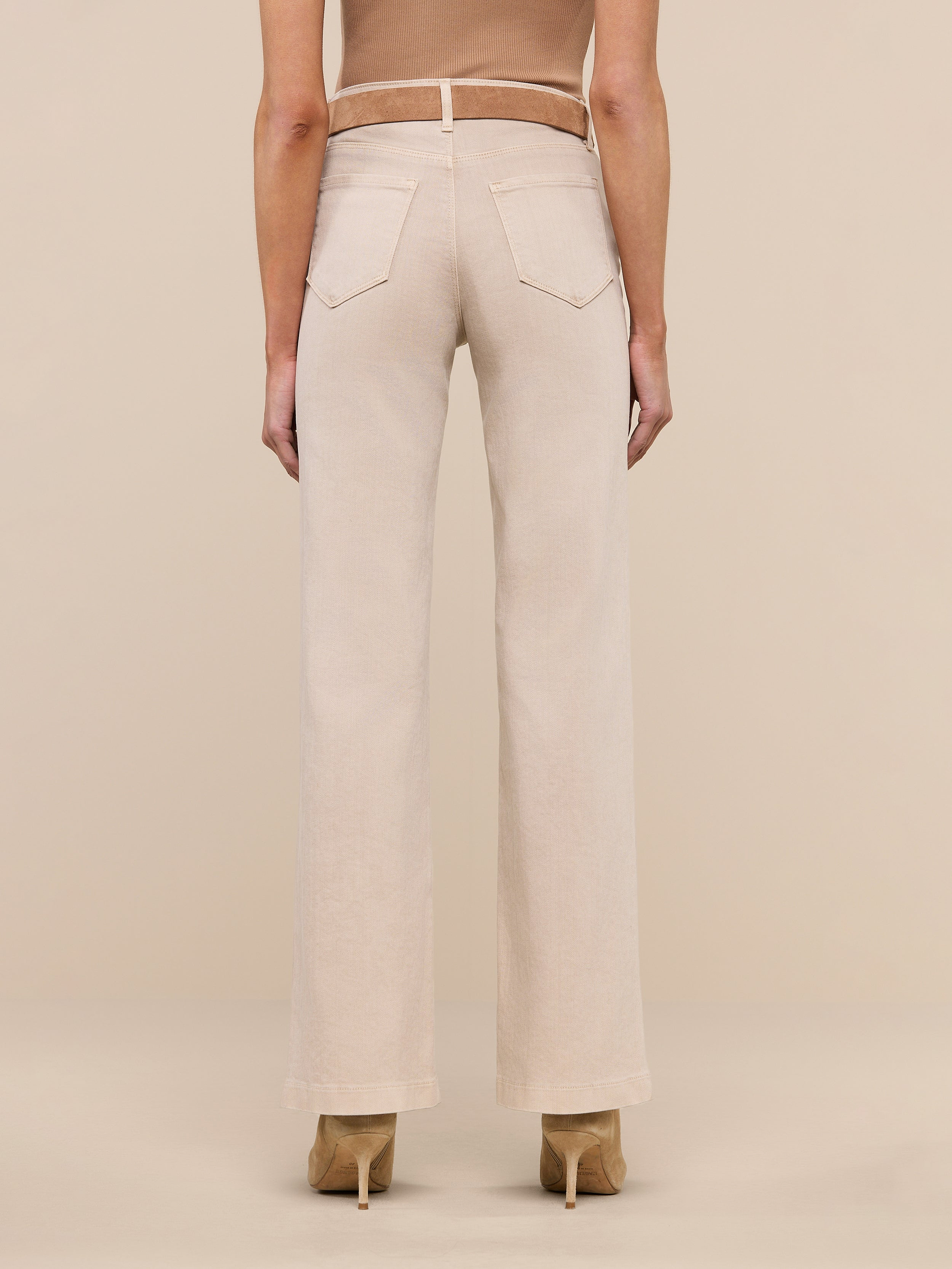 A person stands facing away, wearing the Scottie Wide-Leg Jean in a light beige wash with back pockets and tan high-heeled shoes against a plain white background.