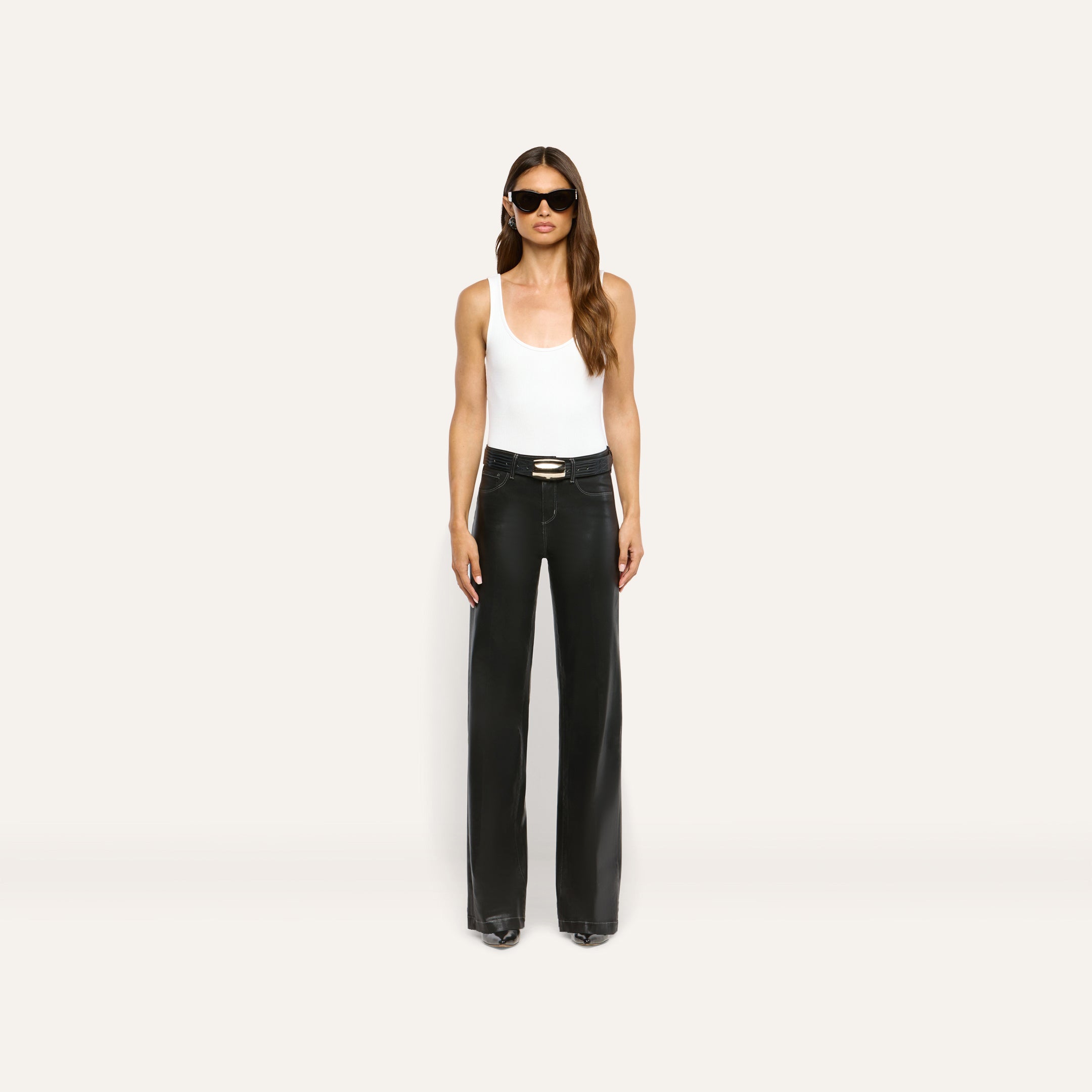 A model wearing a white tank top, a black belt with a gold buckle, and Scottie Coated Wide-Leg Jean high-rise, wide-leg coated denim pants.