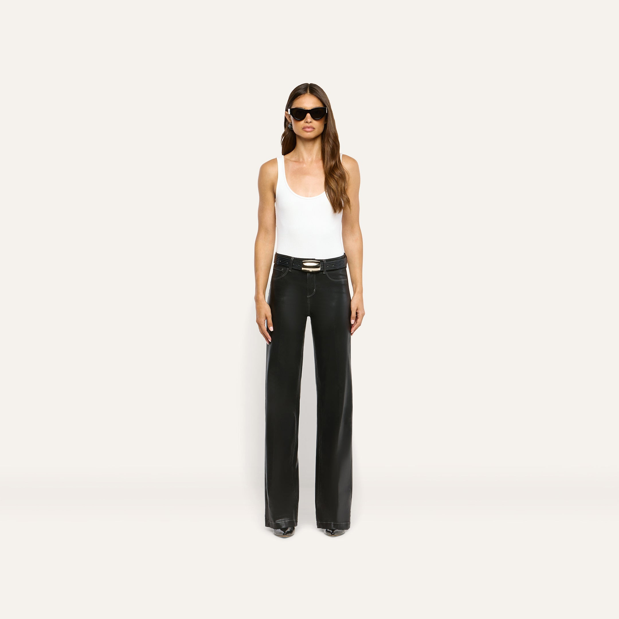 A model wearing a white tank top, a black belt with a gold buckle, and Scottie Coated Wide-Leg Jean high-rise, wide-leg coated denim pants.