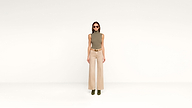 A woman stands against a plain white background, wearing sunglasses, an olive sleeveless top, beige wide-leg pants, a belt, and stylish green ankle boots.