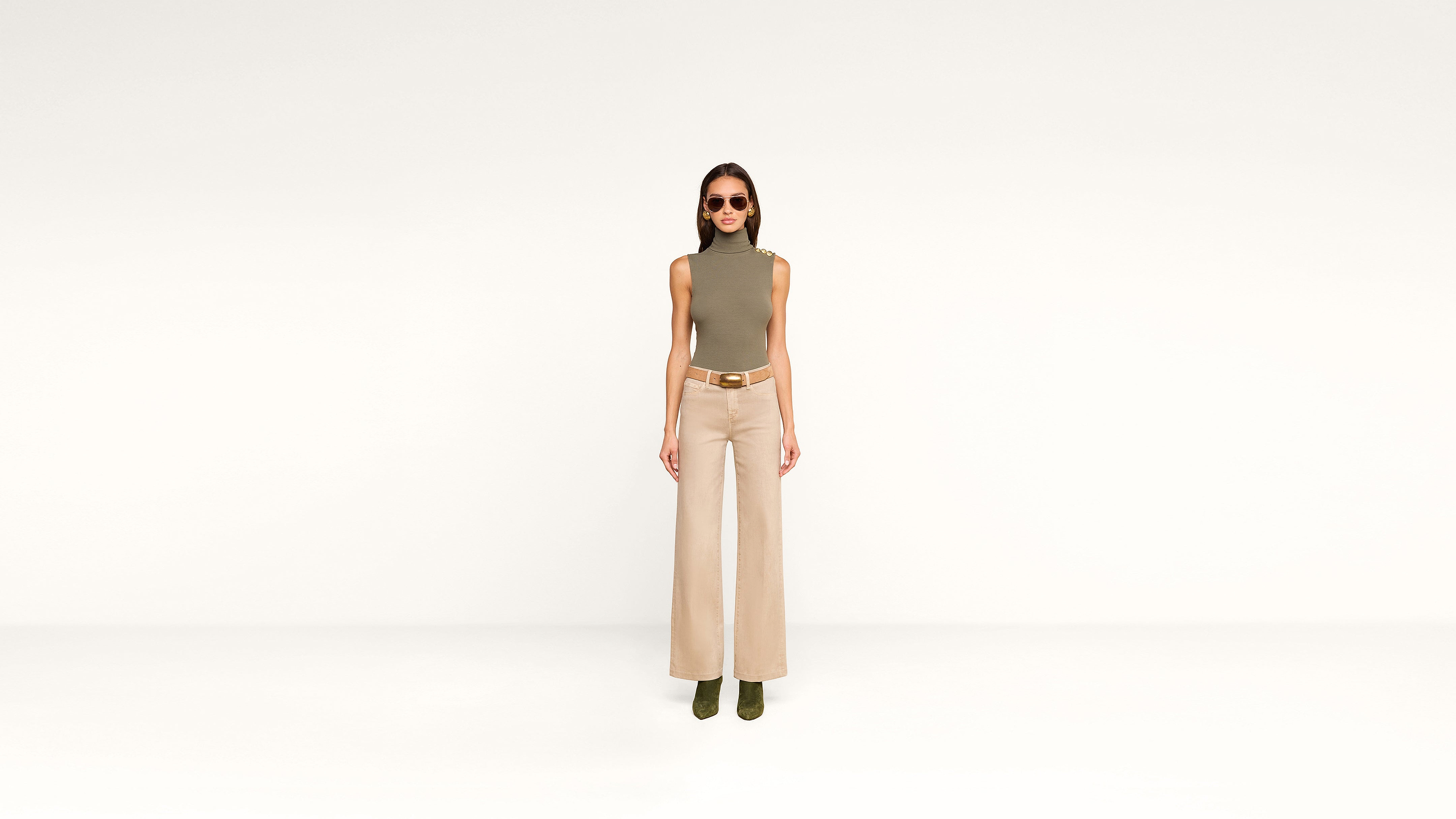 A woman stands against a plain white background, wearing sunglasses, an olive sleeveless top, beige wide-leg pants, a belt, and stylish green ankle boots.