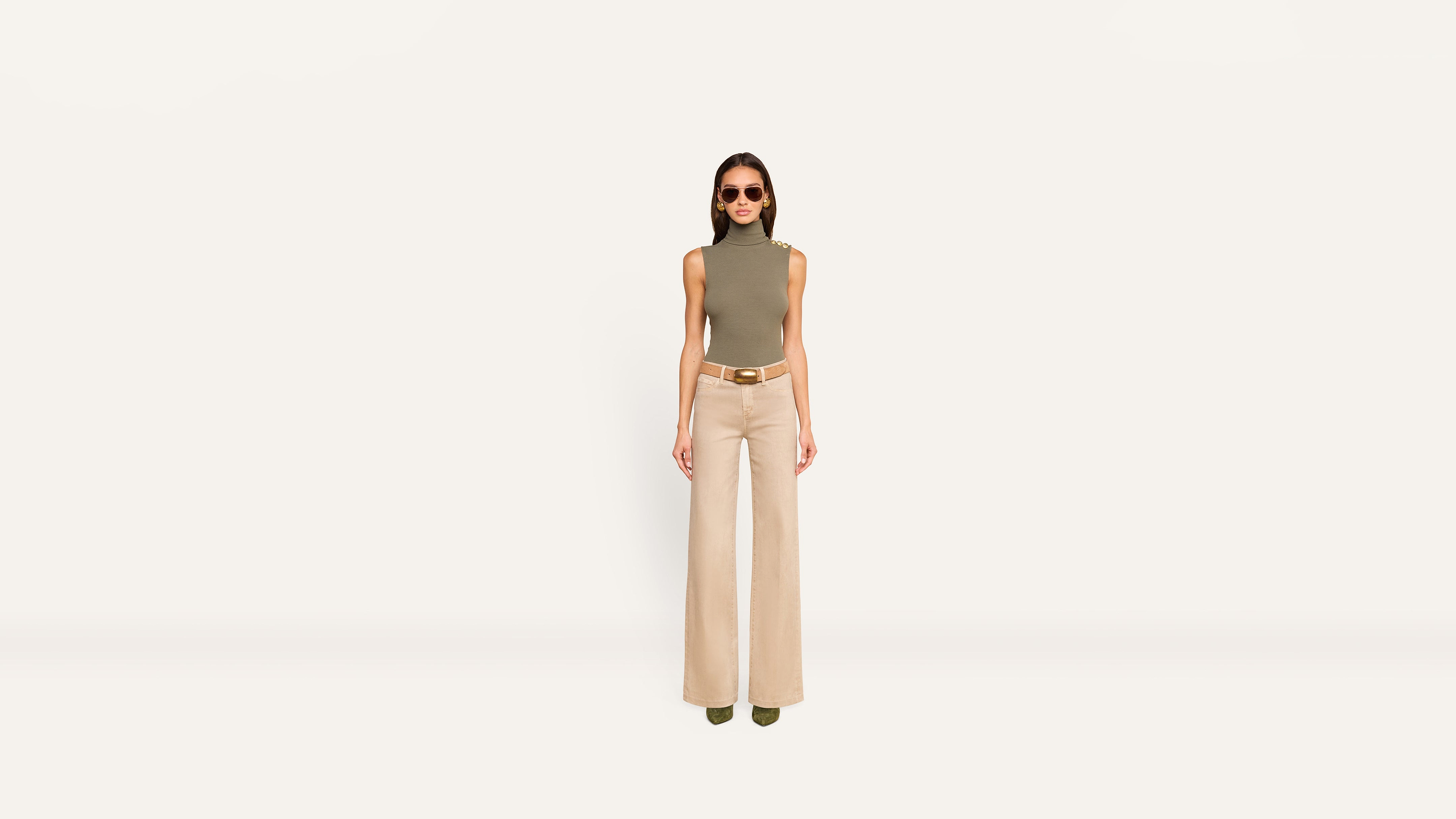 A woman wearing a sleeveless olive turtleneck, a tan belt, and wide-leg beige trousers.