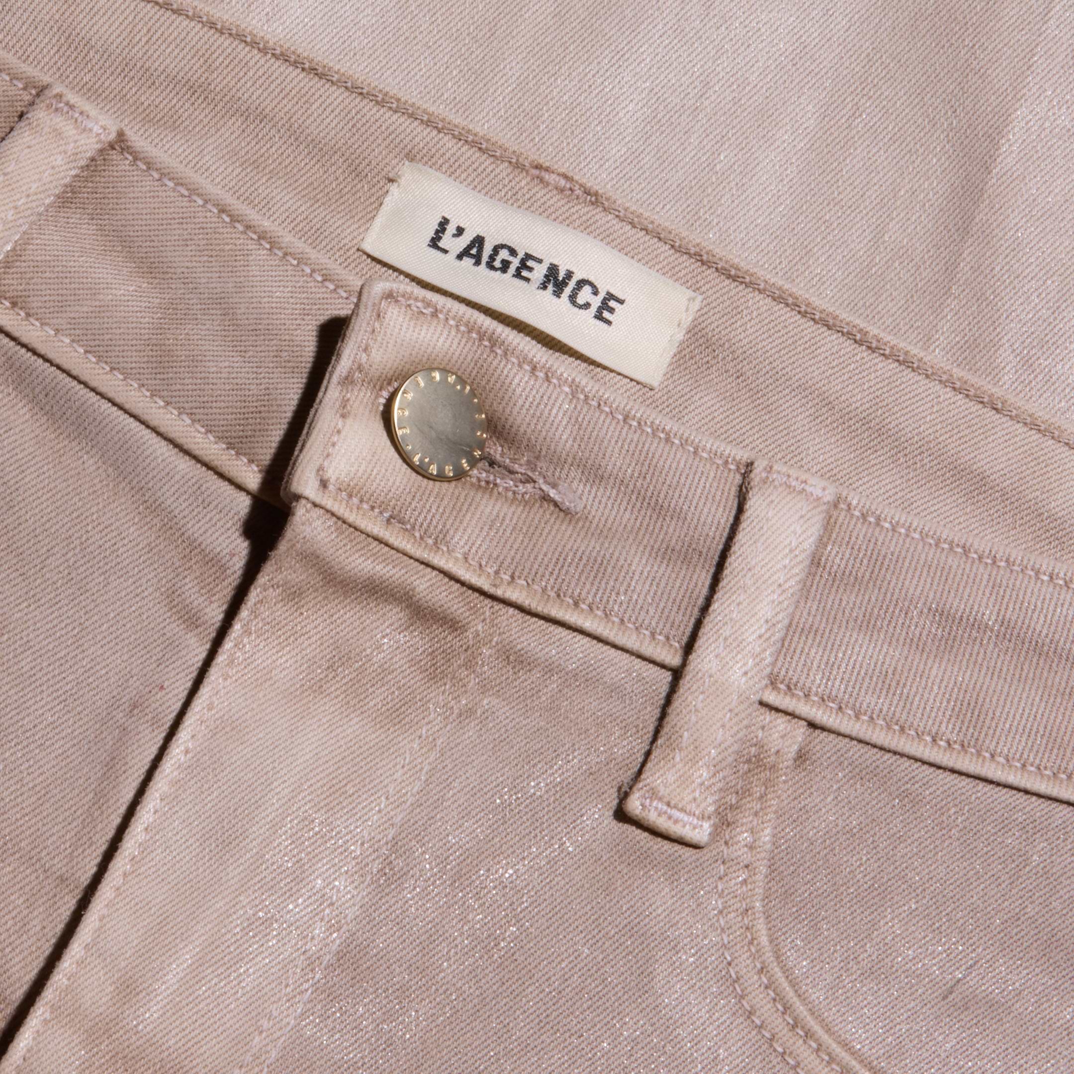 Close-up of the Scottie Coated Wide-Leg Jean in light pink, featuring a metallic button and a white L’AGENCE label sewn onto the waistband. The coated denim fabric gives off a subtle sheen, with visible stitching details.
