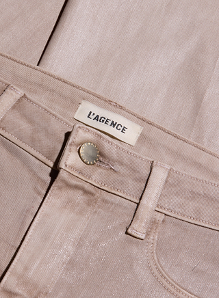Here’s a revised version using the provided product name: Close-up of the Scottie Coated Wide-Leg Jean in light pink, highlighting a metallic button, front pocket, belt loop, and a white tag with LAGENCE in bold black letters. These high-rise jeans are folded neatly against a matching fabric background.