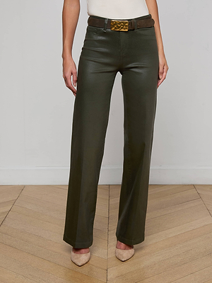 A person wears the L'AGENCE Scottie Coated Wide-Leg Jean in dark green, paired with a brown belt with a gold buckle, a white top, and beige pointed-toe heels, standing on a wooden floor against a white wall.