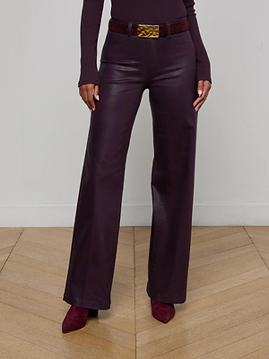 A person stands indoors wearing L'AGENCE Scottie Coated Wide-Leg Jeans in dark burgundy, a matching fitted top, pointed burgundy shoes, and a dark belt with a gold buckle; only their body from shoulders down is visible.