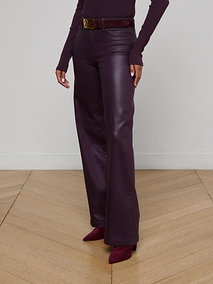 A person stands on a wooden floor wearing L’AGENCE’s Scottie Coated Wide-Leg Jean in dark purple with a matching long-sleeve top, wide belt, and pointed-toe heeled boots; their face is not visible.