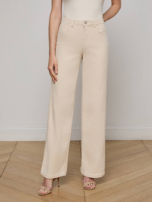 A person wears the L'AGENCE Scottie Corduroy Wide-Leg Pant in high-waisted stretch corduroy, paired with a white sleeveless top and nude heeled sandals, standing on a wooden floor against a plain white wall.