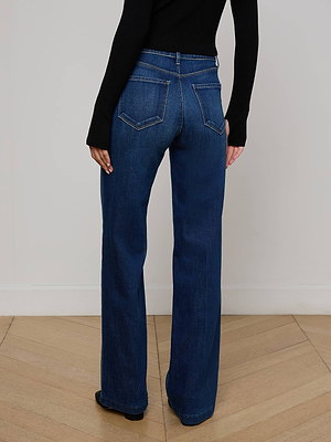 A person stands with their back to the camera on a light wooden floor, wearing L'AGENCE Scottie Wide-Leg Jeans in dark blue with a contoured waistband and a fitted black long-sleeve top.