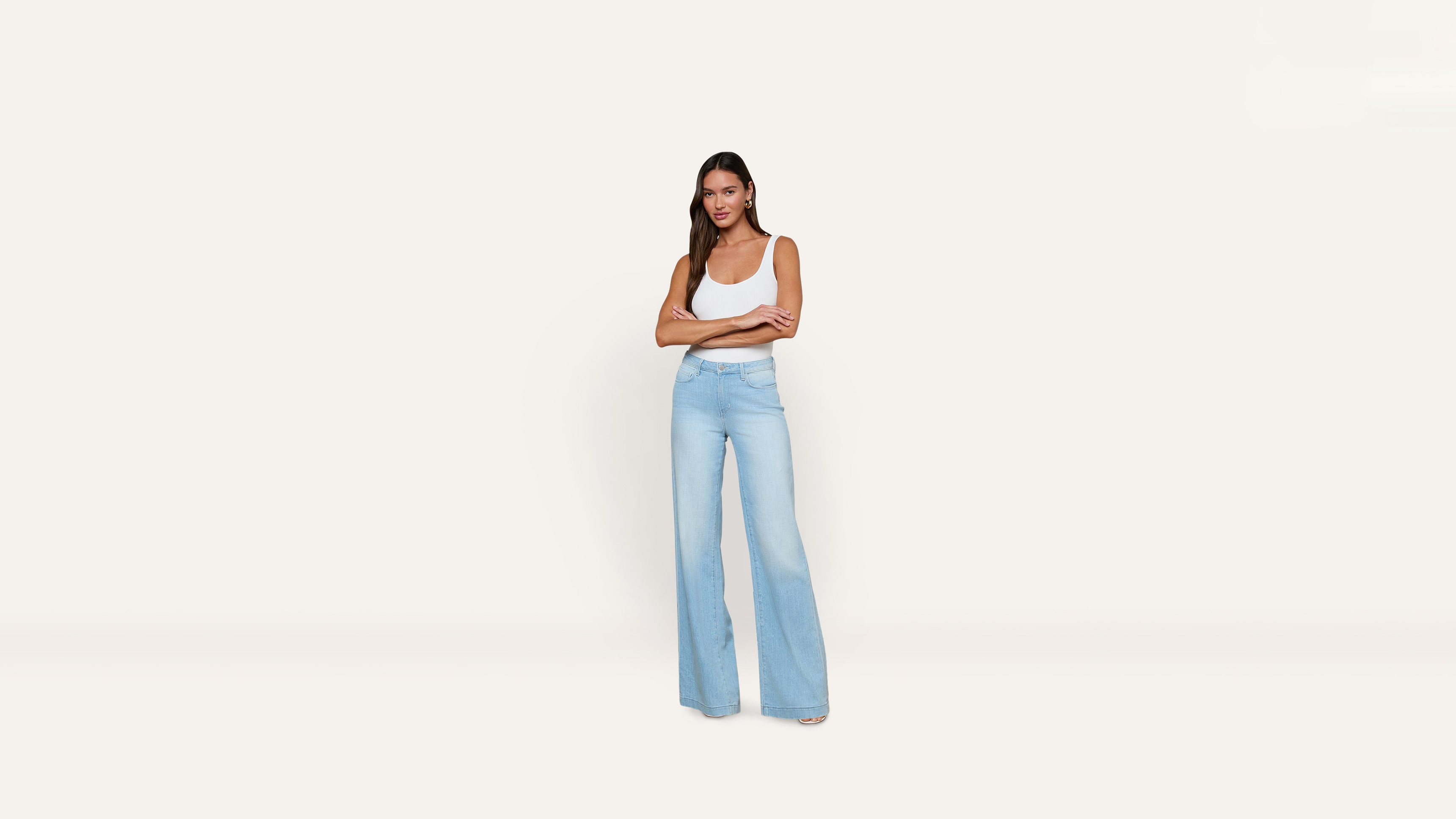 A woman stands with her arms crossed, wearing a white tank top and light-wash wide-leg jeans.