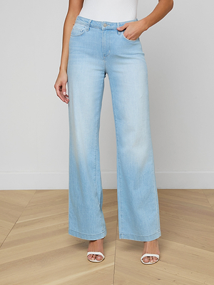 Someone wearing the Scottie Wide-Leg Jean in light blue with a contoured waistband, paired with a white top and white heeled sandals, stands on a light wooden floor against a plain white wall.