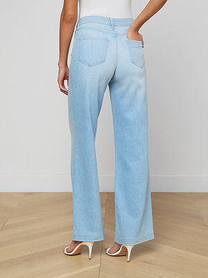 Person shown from the back standing on a light wooden floor, wearing Scottie Wide-Leg Jean in light blue with a contoured waistband, a white top, and white heels; one hand rests in the jeans’ back pocket.