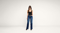 A woman wearing sunglasses, a black tank top, blue high-waisted jeans, and black heels stands against a plain white background.