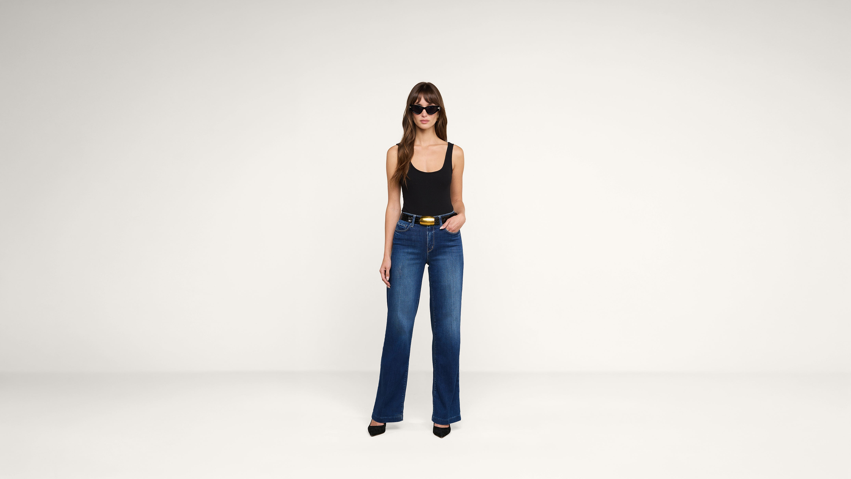 A woman wearing sunglasses, a black tank top, blue high-waisted jeans, and black heels stands against a plain white background.