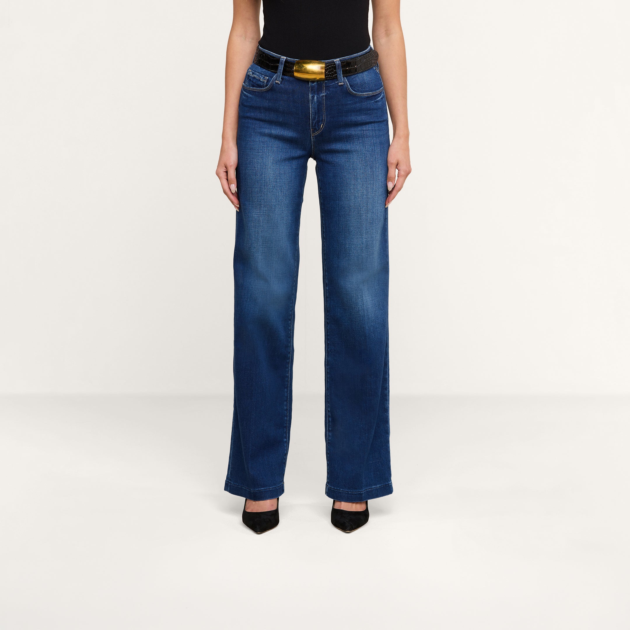 A person, visible from the shoulders down against a plain background, is wearing a black sleeveless top, the Scottie Wide-Leg Jean in blue stretch denim with a high waist, styled with a black and gold belt and paired with black pointed-toe heels.