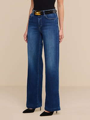 A person stands against a plain beige background wearing Scottie Wide-Leg Jeans with a contoured waistband, styled with a black belt and black pointed-toe heels.