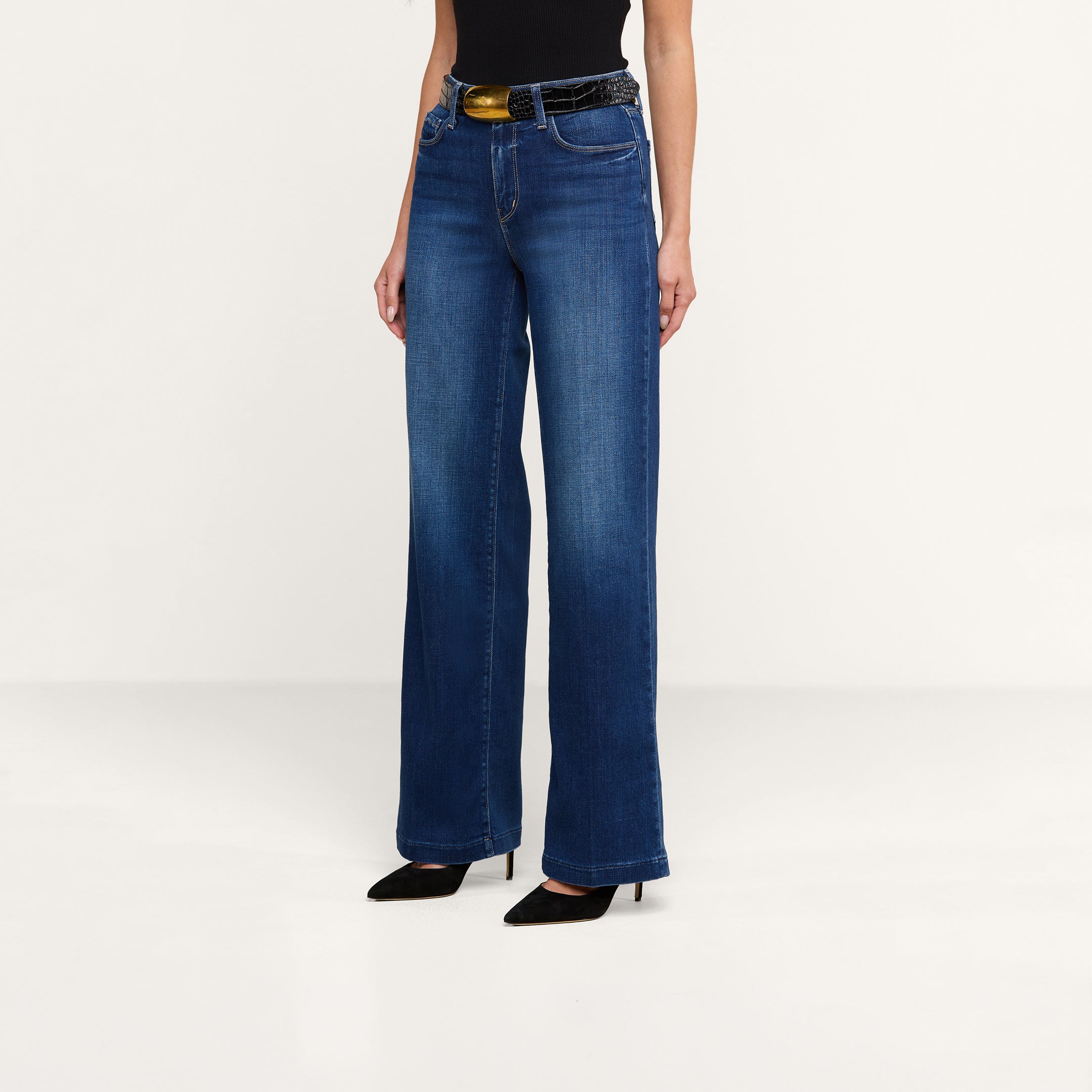 A person wearing the Scottie Wide-Leg Jean with a contoured waistband, paired with a black belt featuring a gold buckle, a black sleeveless top, and black pointed-toe heels stands against a plain white background.