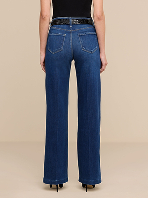 A person stands with their back to the camera against a beige background, wearing Scottie Wide-Leg Jeans in blue, a contoured waistband, a black belt, and a sleeveless black top.