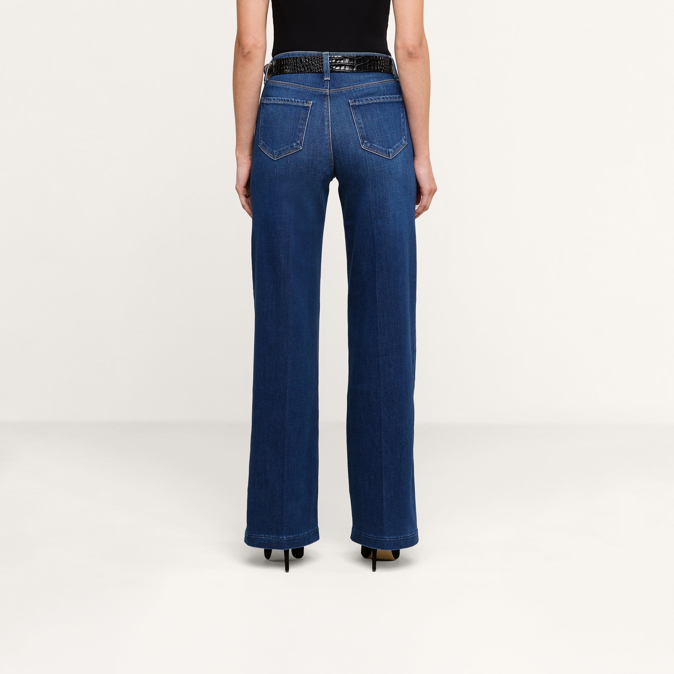 Wearing the Scottie Wide-Leg Jean in dark blue with a contoured waistband and back pockets, paired with a black sleeveless top, black belt, and black heeled shoes, a person stands facing away against a plain white background.