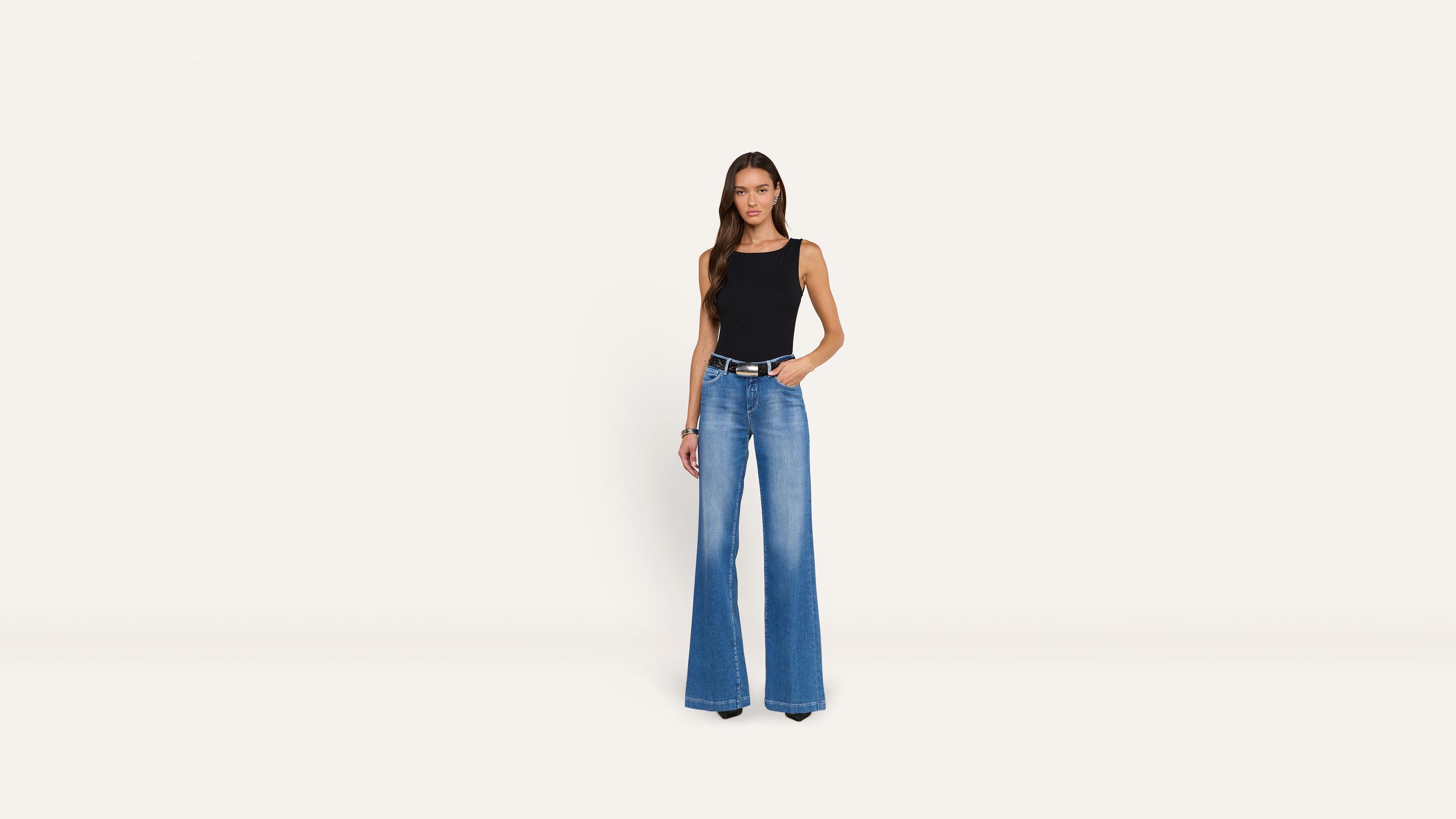 A woman stands against a plain background wearing a black sleeveless top and blue wide-leg jeans cinched with a black belt.