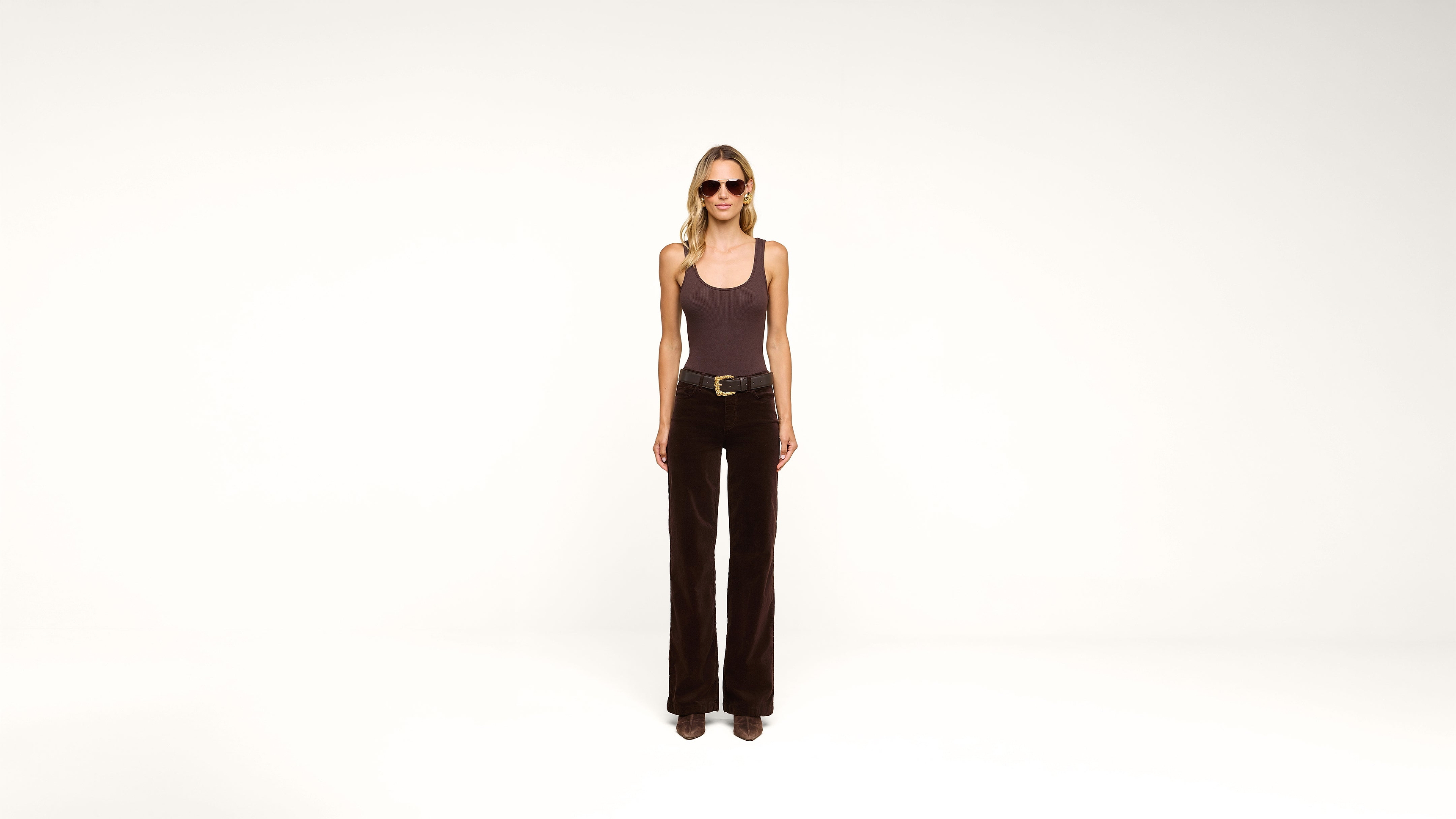A person wearing sunglasses, a dark tank top, dark pants, and boots stands against a plain white background, facing forward with hands relaxed at their sides.
