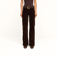 A person stands against a plain white background, wearing the Scottie Velvet Wide-Leg Jean in dark brown with a contoured waistband, paired with a black belt and brown pointed shoes.