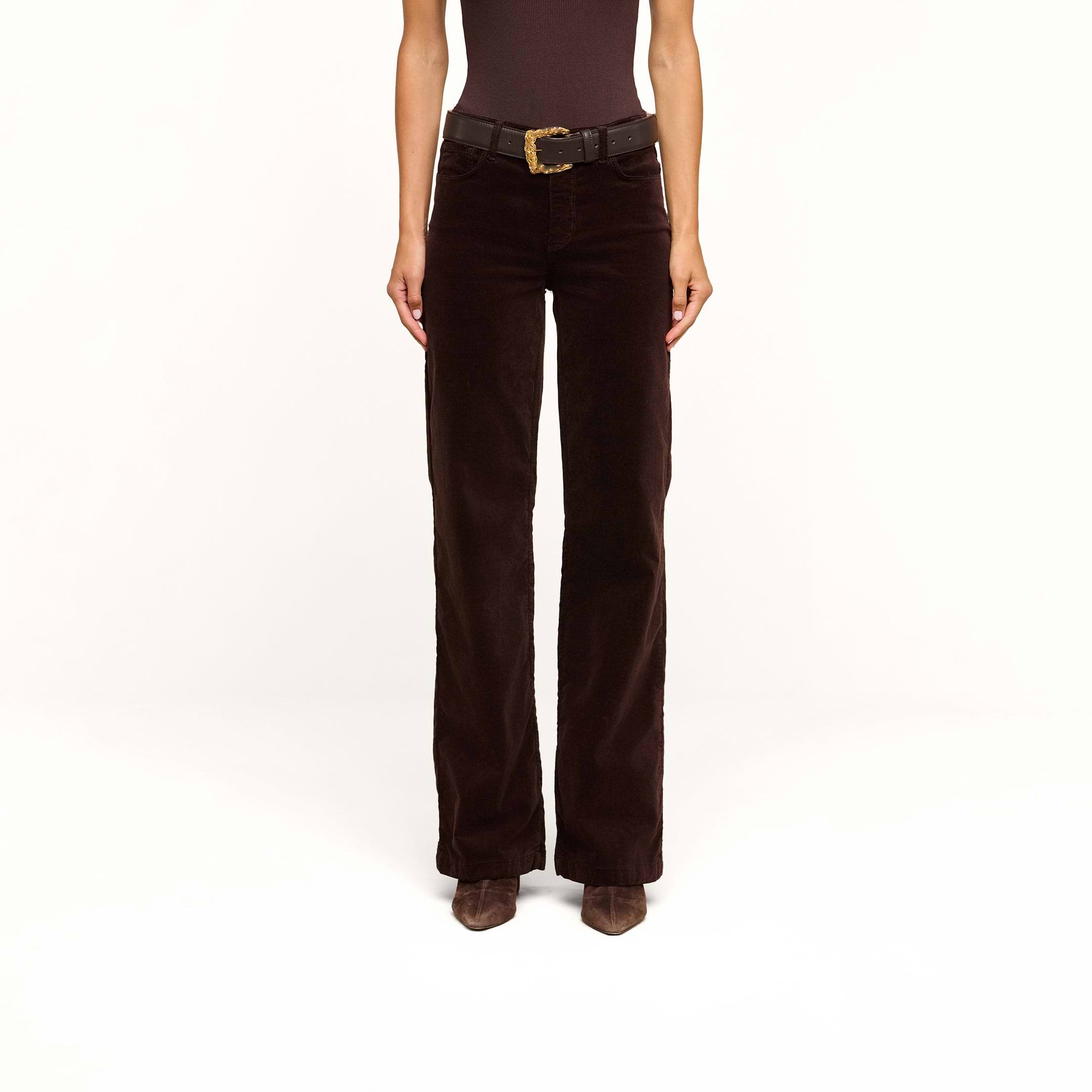 A person stands against a plain white background, wearing the Scottie Velvet Wide-Leg Jean in dark brown with a contoured waistband, paired with a black belt and brown pointed shoes.