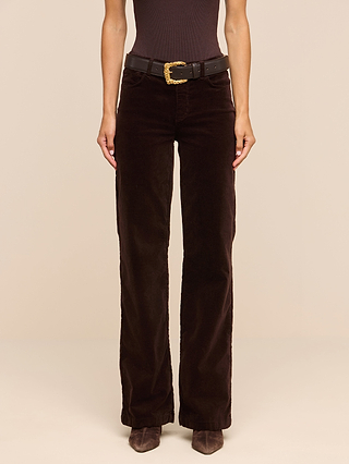 Person wearing a black top, the Scottie Velvet Wide-Leg Jean in high-rise dark brown, a black belt with a gold buckle, and brown shoes, standing against a plain beige background—an effortlessly stylish look perfect for any transitional wardrobe.