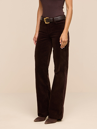 Woman wearing Scottie Velvet Wide-Leg Jeans in dark brown, styled with a black belt featuring a gold buckle, a sleeveless brown top, and brown shoes, standing against a plain beige background. Her arms are relaxed by her sides—a perfect look for building your transitional wardrobe.