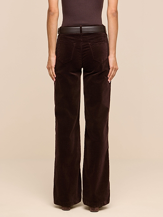 A person stands facing away, wearing the Scottie Velvet Wide-Leg Jean in dark brown with a matching belt and a sleeveless top—an effortlessly chic choice for any transitional wardrobe. The background is plain beige.