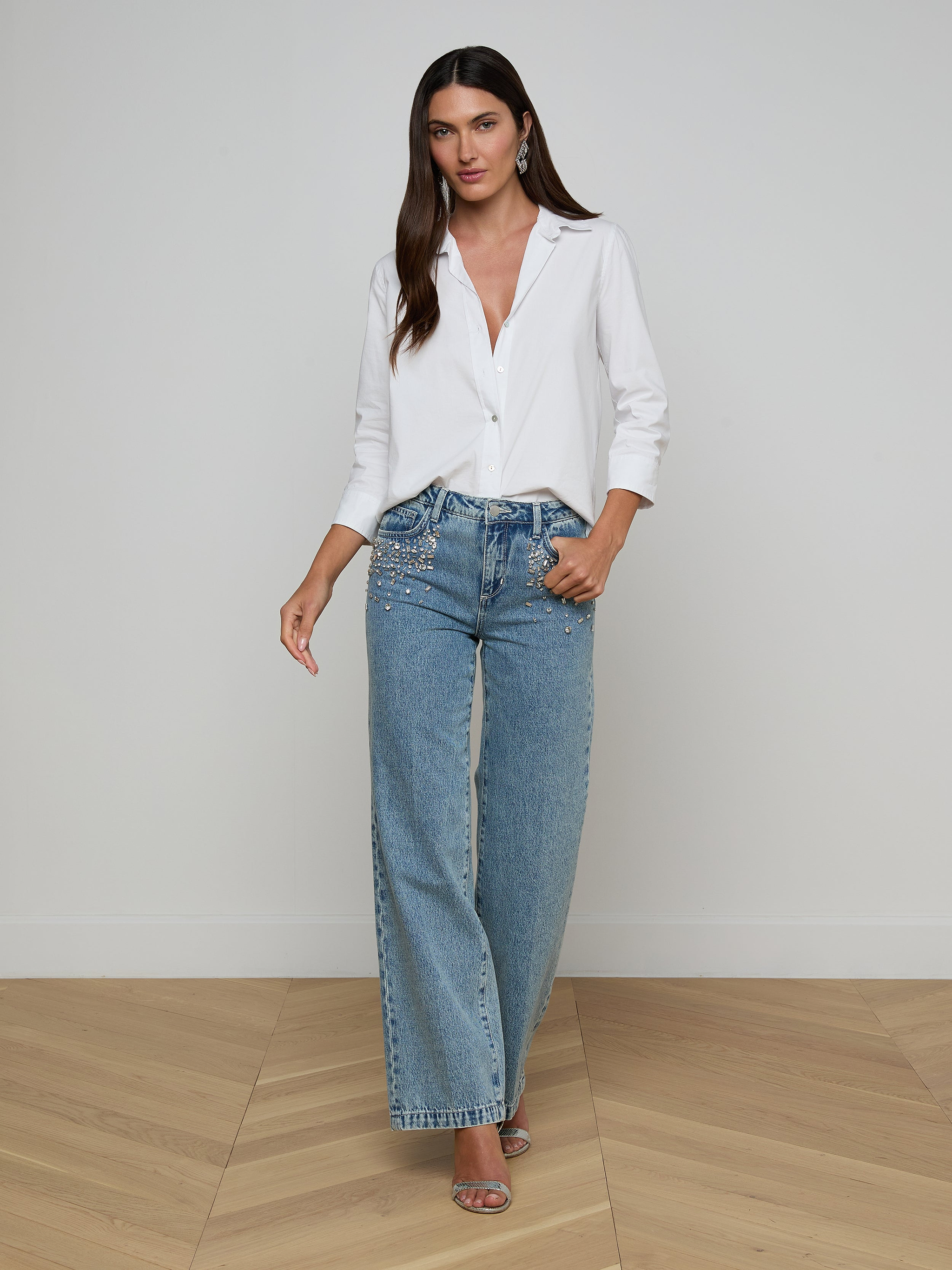 A woman stands against a plain white background, wearing a white button-up shirt with rolled sleeves, the Scottie Embellished Wide-Leg Jean—crafted from all-cotton denim in a high-waisted, wide-leg silhouette—and open-toe heels. She has long, straight hair and a neutral expression.