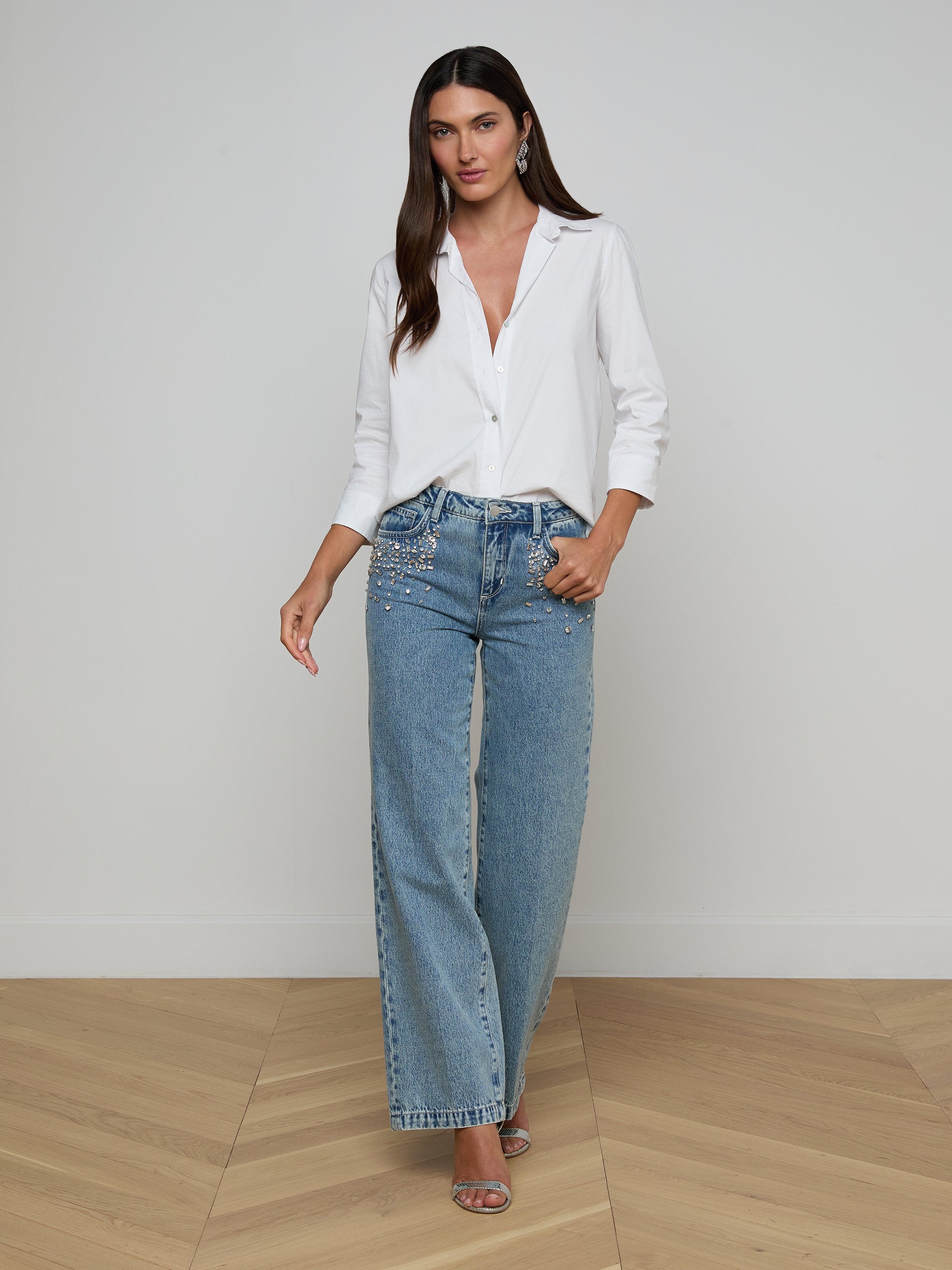 A woman stands against a plain white background, wearing a white button-up shirt with rolled sleeves, the Scottie Embellished Wide-Leg Jean—crafted from all-cotton denim in a high-waisted, wide-leg silhouette—and open-toe heels. She has long, straight hair and a neutral expression.