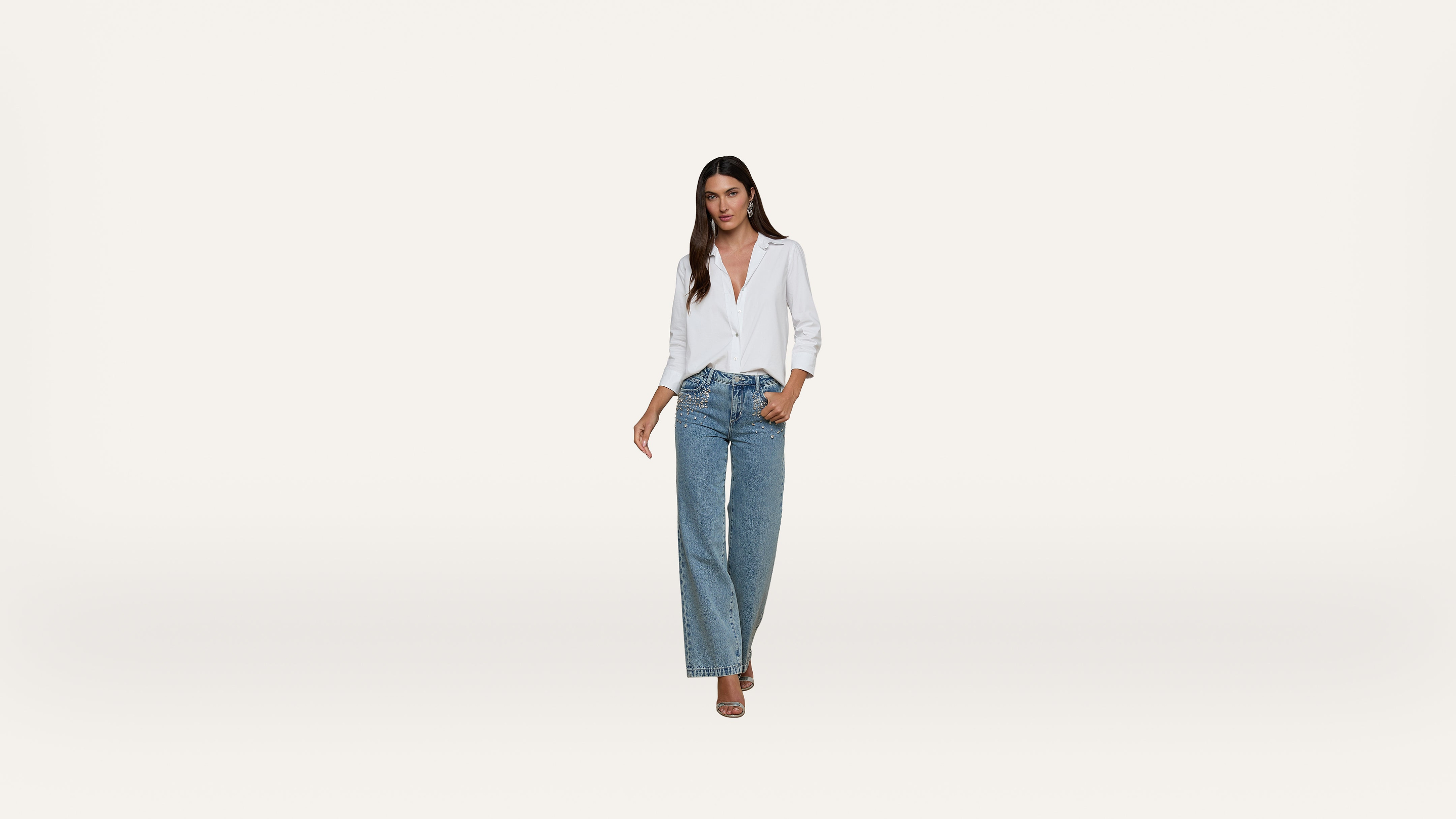A woman wearing a white blouse and wide-leg blue jeans stands against a plain white background, looking confidently at the camera.