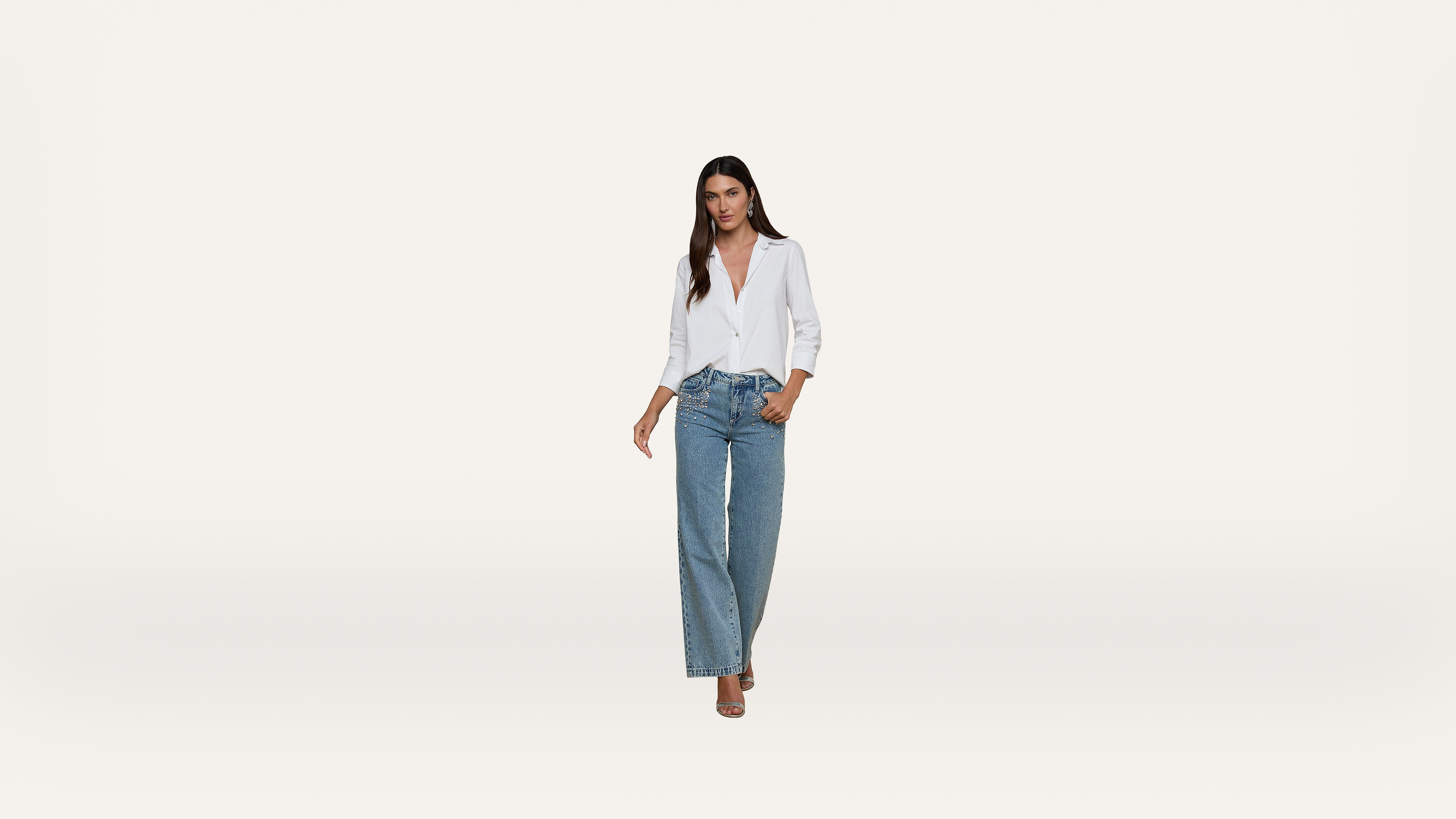 A woman wearing a white blouse and wide-leg blue jeans stands against a plain white background, looking confidently at the camera.