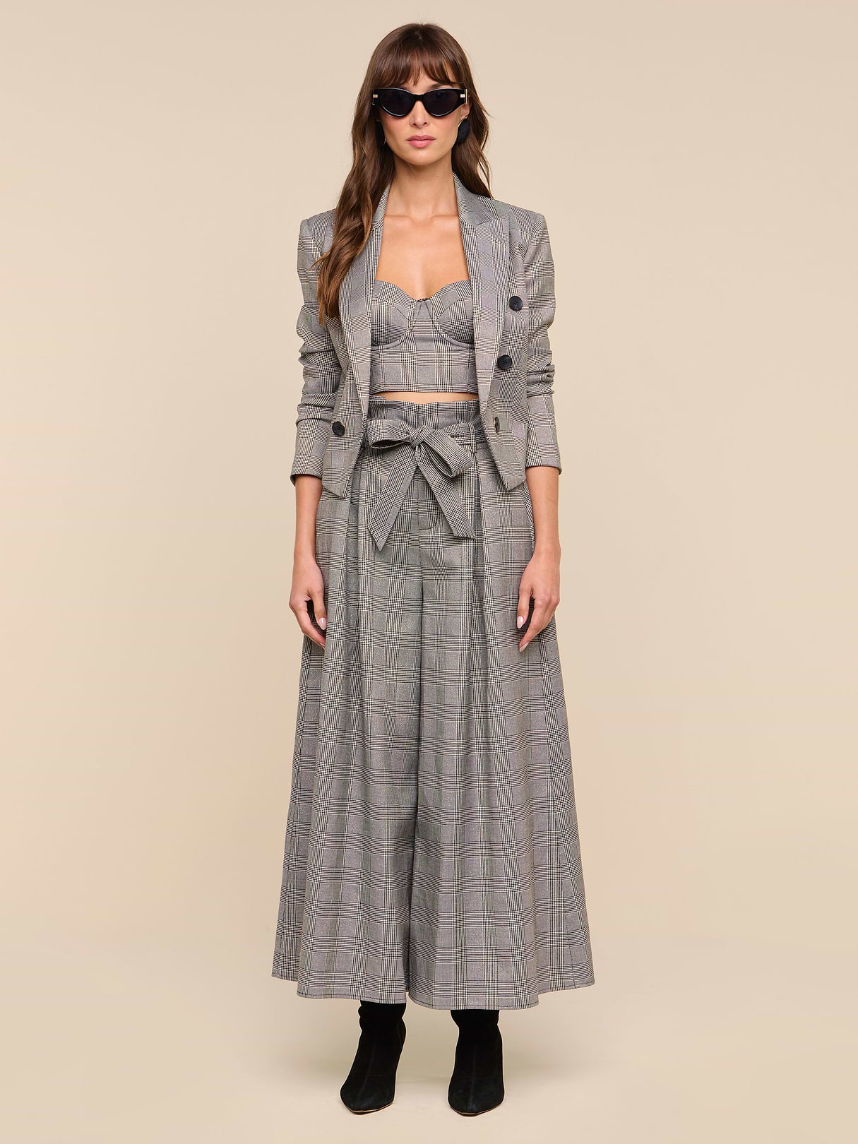 A woman faces forward against a plain white background, wearing the Lowen Pant—a pair of high-rise, wide-leg gray plaid trousers—styled with a matching cropped blazer, bralette, and black boots. She has long hair, sunglasses, and a neutral expression.
