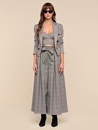 A woman faces forward against a plain white background, wearing the Lowen Pant—a pair of high-rise, wide-leg gray plaid trousers—styled with a matching cropped blazer, bralette, and black boots. She has long hair, sunglasses, and a neutral expression.