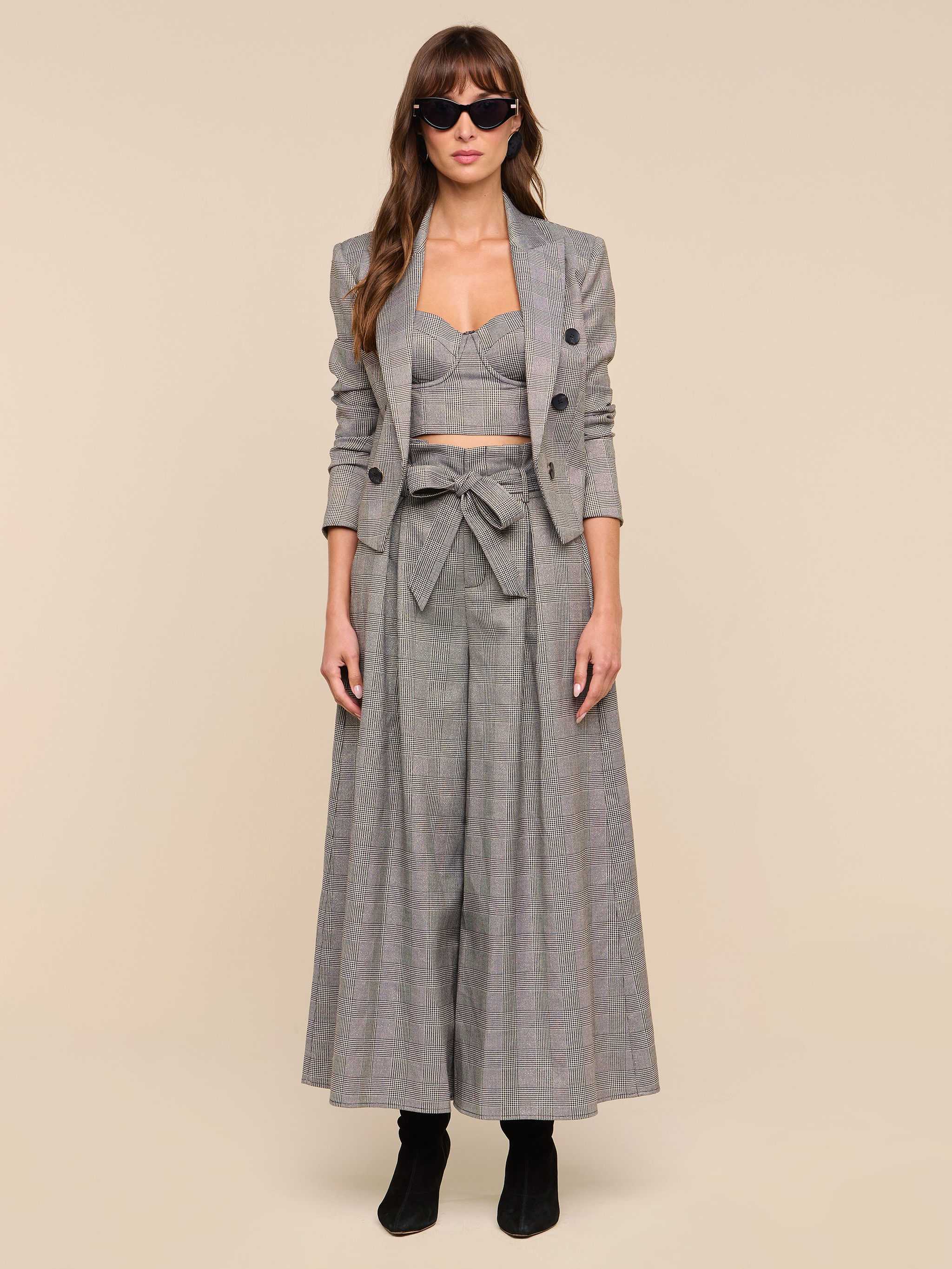 A woman faces forward against a plain white background, wearing the Lowen Pant—a pair of high-rise, wide-leg gray plaid trousers—styled with a matching cropped blazer, bralette, and black boots. She has long hair, sunglasses, and a neutral expression.
