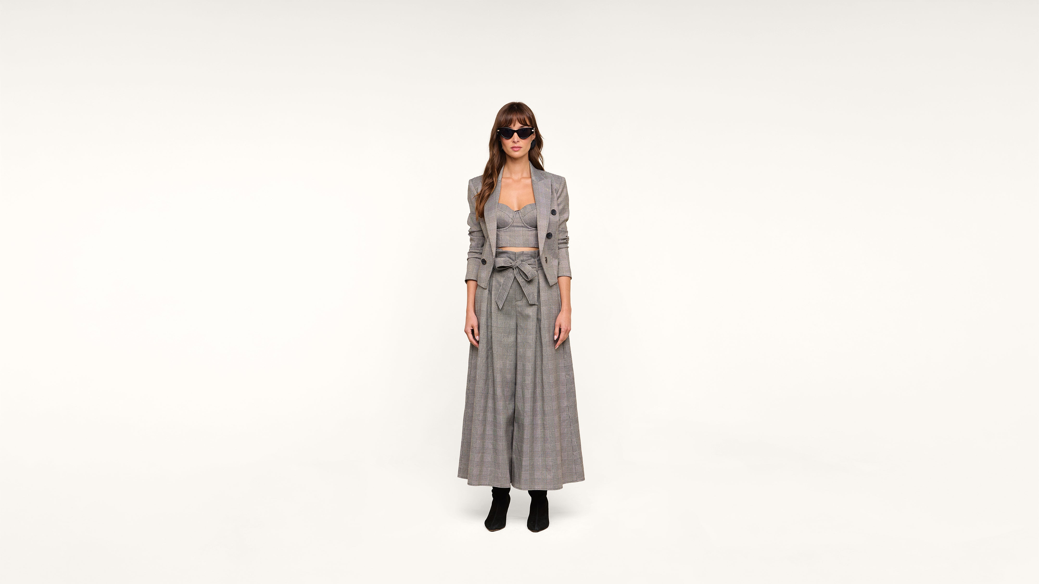 A woman wearing sunglasses, a gray plaid blazer, and a matching long skirt stands facing forward against a plain white background. She is also wearing black ankle boots.