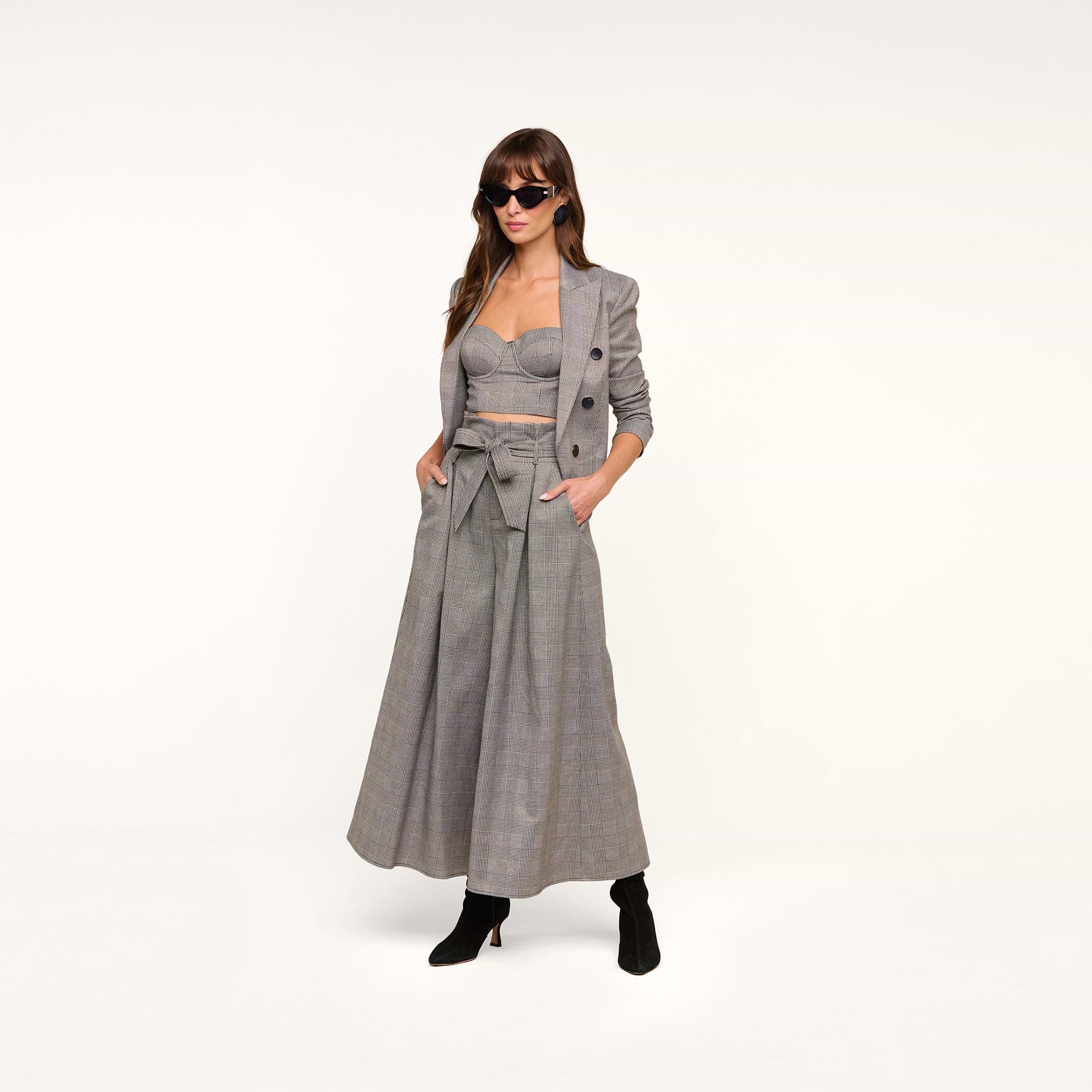 Woman wearing sunglasses, a gray plaid suiting blazer, cropped bustier top, and the Lowen Pant—high-rise, wide-leg pants featuring a paper-bag waist with a bow belt—paired with black ankle boots, standing against a plain white background.