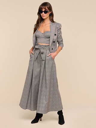 Woman wearing the Lowen Pant—a pair of high-rise, wide-leg pants with a paper-bag waist and bow belt—in gray plaid, paired with a matching blazer and bralette, black boots, and sunglasses, standing with hands in pockets against a beige background.