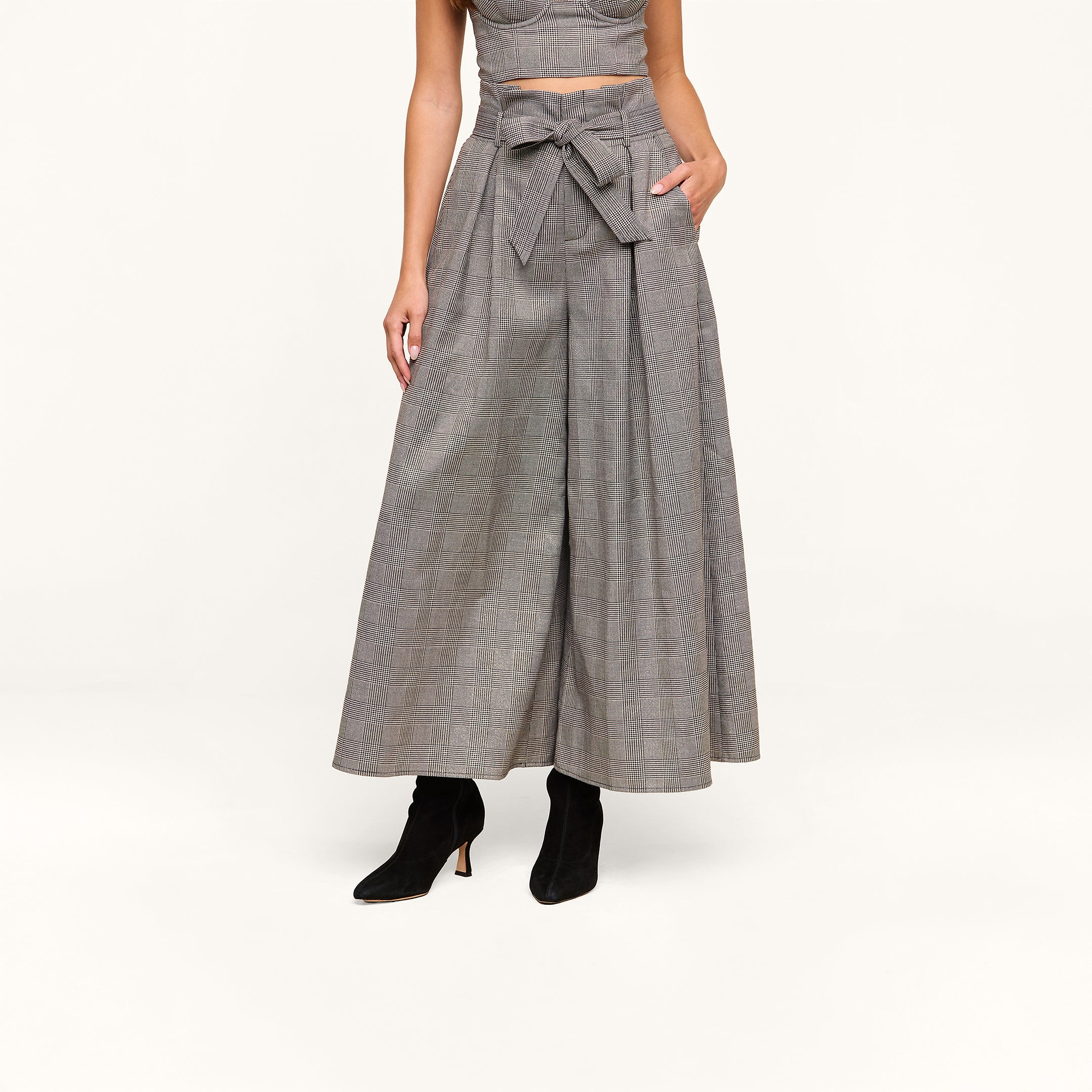 A person wearing the Lowen Pant—a pair of wide-leg, high-rise plaid suiting culottes with a matching belt—styled with black heeled ankle boots and a sleeveless crop top. Their face is not visible.
