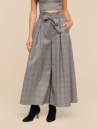 A person wearing the Lowen Pant, a wide-leg, high-rise gray plaid suiting pant with a matching belt, along with a sleeveless top and black heeled boots, stands against a plain beige background.