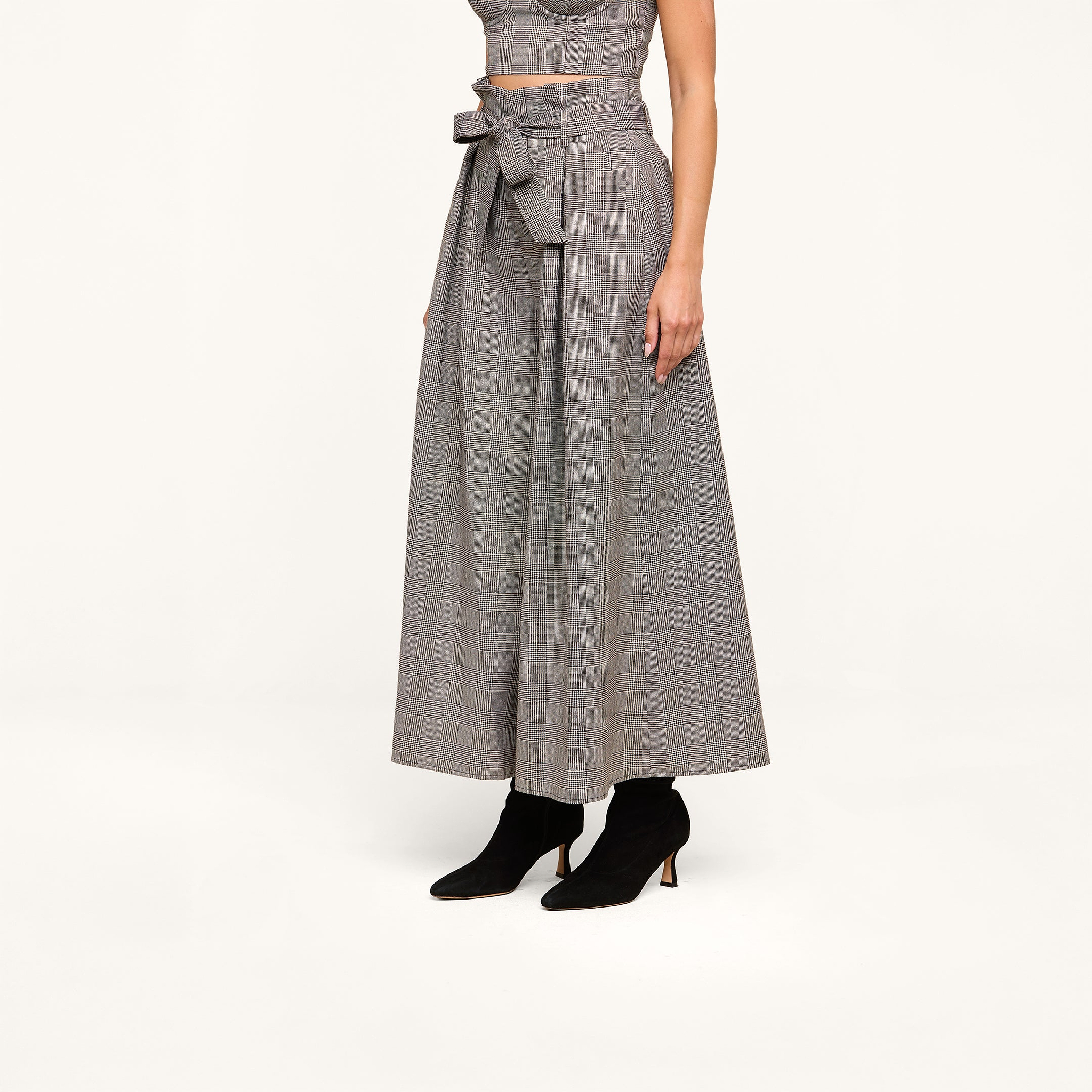 A person wearing the Lowen Pant—high rise, wide-leg gray plaid suiting pants with a paper-bag waist tie—paired with black heeled ankle boots and a matching sleeveless crop top poses against a plain white background.