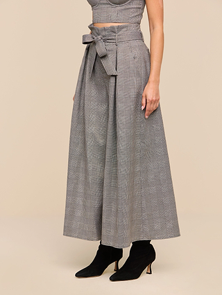 A woman wearing the Lowen Pant—high-waisted, wide-leg gray plaid suiting pants featuring a paper-bag waist with a matching belt tied in a bow—paired with a sleeveless top and black heeled ankle boots.