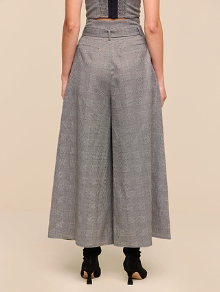 A person is shown from the back, standing against a plain beige background and wearing the Lowen Pant—wide-leg gray plaid suiting trousers—paired with black heeled shoes.